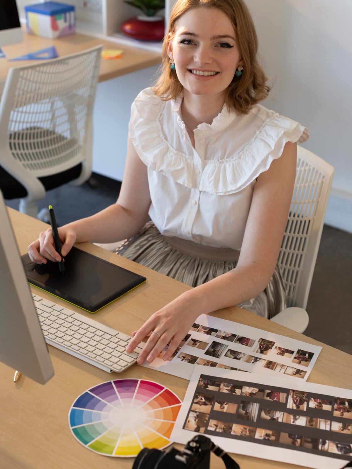 Portrait of young Caucasian female graphic designer working on graphics tablet and computer at desk in the office. She is smiling and looking at camera